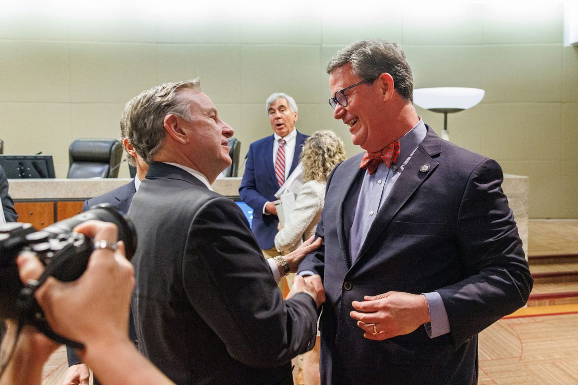 Alt text: Two men in suits shake hands in a formal chamber, with a photographer in the foreground and several people standing and talking in the background. The setting appears to be an official meeting or ceremonial event.