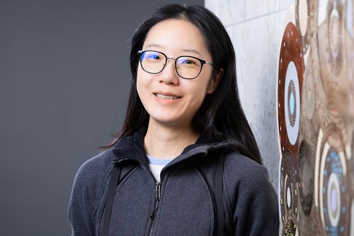 A woman with long black hair and glasses is smiling while leaning against a patterned wall.