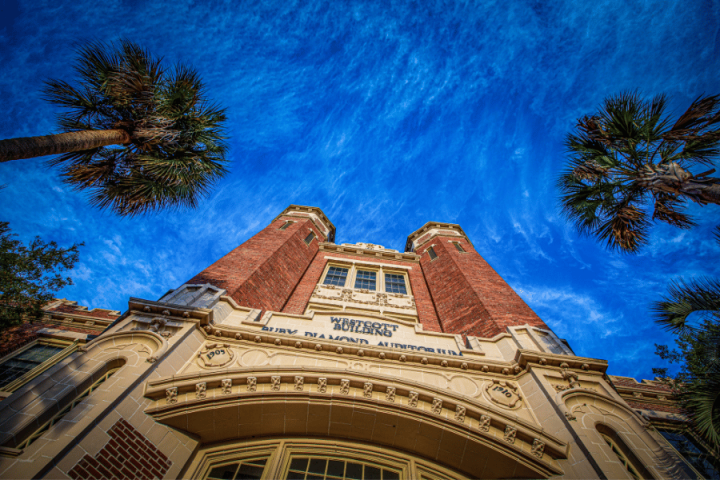 A low-angle view looks up at the ornate brick facade of the Westcott Building and Ruby Diamond Auditorium framed by palm trees against a dramatic blue sky.