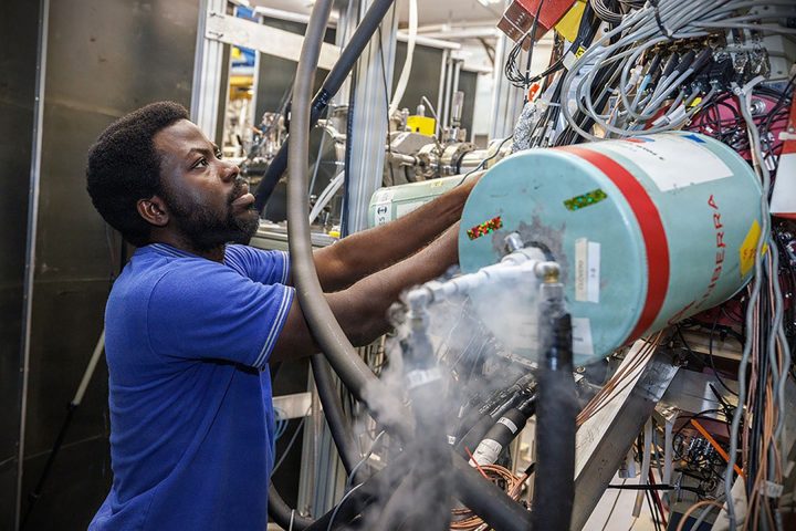 A man works on a large piece of laboratory equipment surrounded by complex wiring and a small amount of vapor rising from a connection.