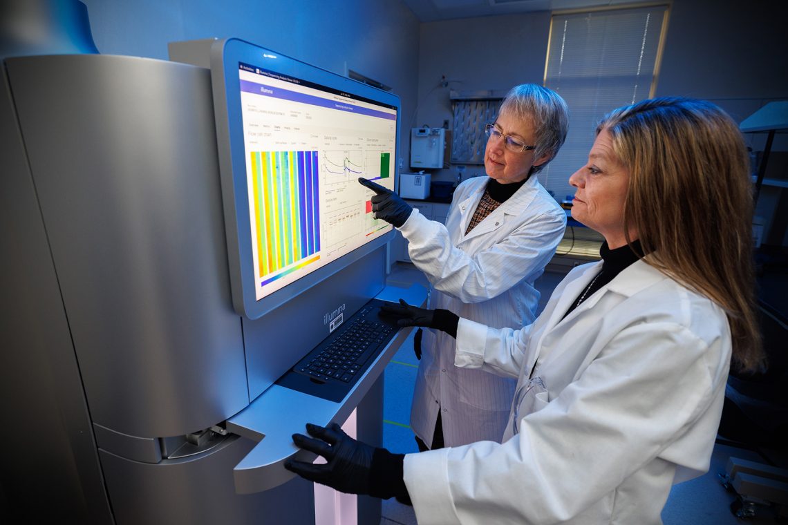 Two scientists wearing white lab coats and black gloves analyze data together on a large, bright touchscreen interface of an Illumina gene sequencing machine in a laboratory environment.