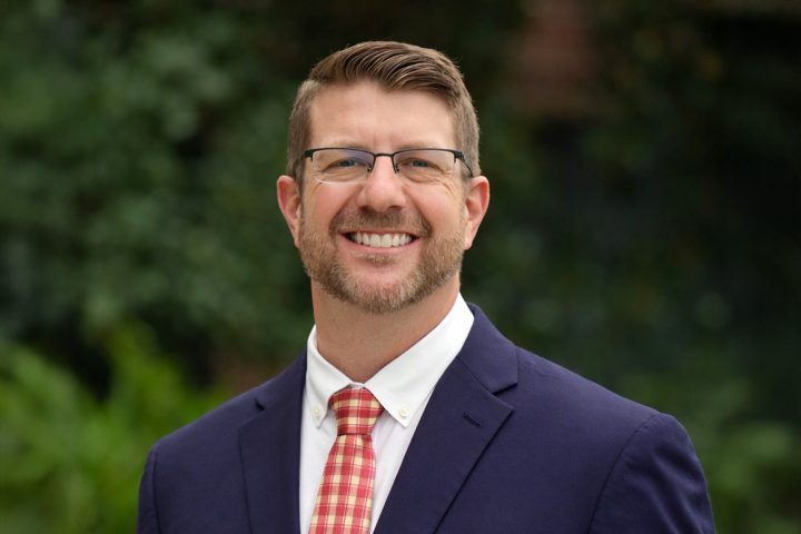 A professional headshot of a smiling man with glasses, light brown hair, and a trimmed beard, wearing a navy blue suit jacket, a white dress shirt, and a red checkered tie against a blurred green background.