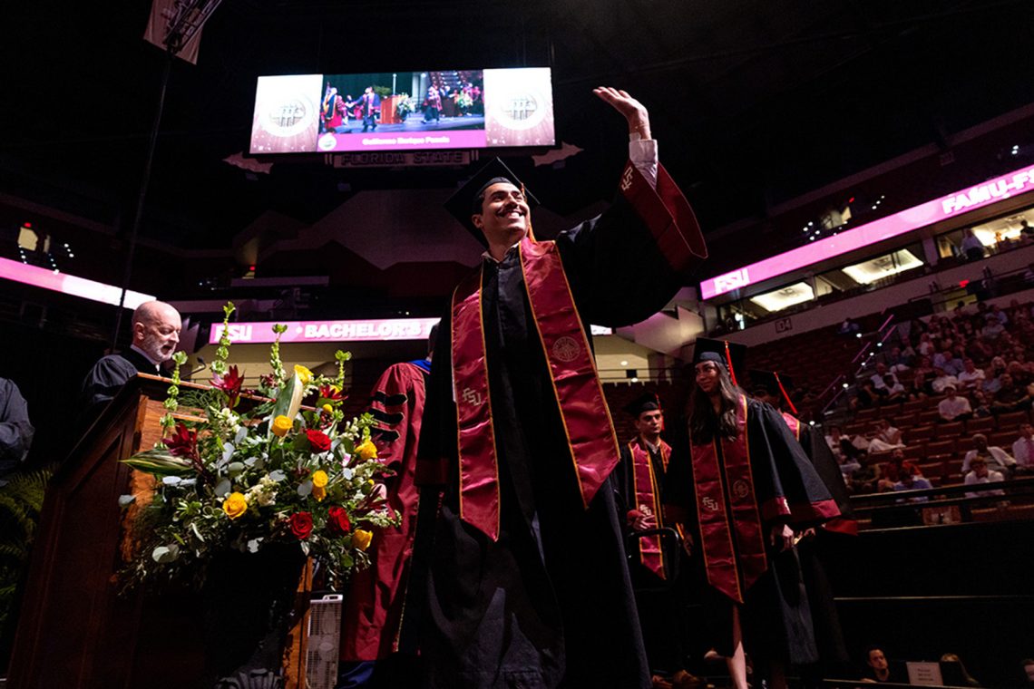 A smiling graduate waves to the audience while walking across the stage during a graduation ceremony.
