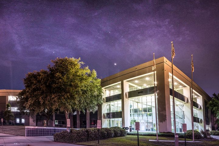 A multi-story modern office building illuminated at night under a purple, starry sky, with flags standing in front.