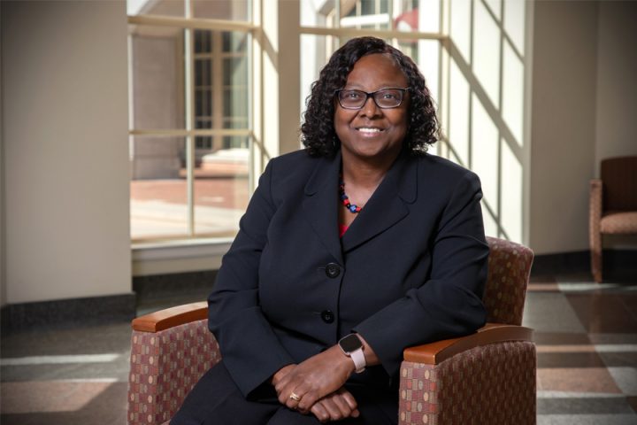 A smiling woman wearing glasses and a black blazer sits in an armchair inside a brightly lit building.