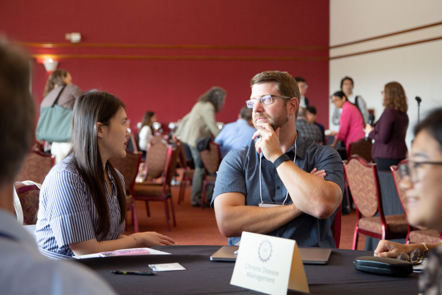 Two attendees converse at a table during a networking event, with other participants mingling in the background