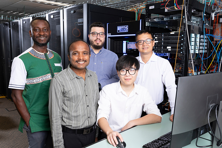 Five researchers stand together in a server room beside a desktop computer, with server racks, networking equipment, and cabling visible in the background.