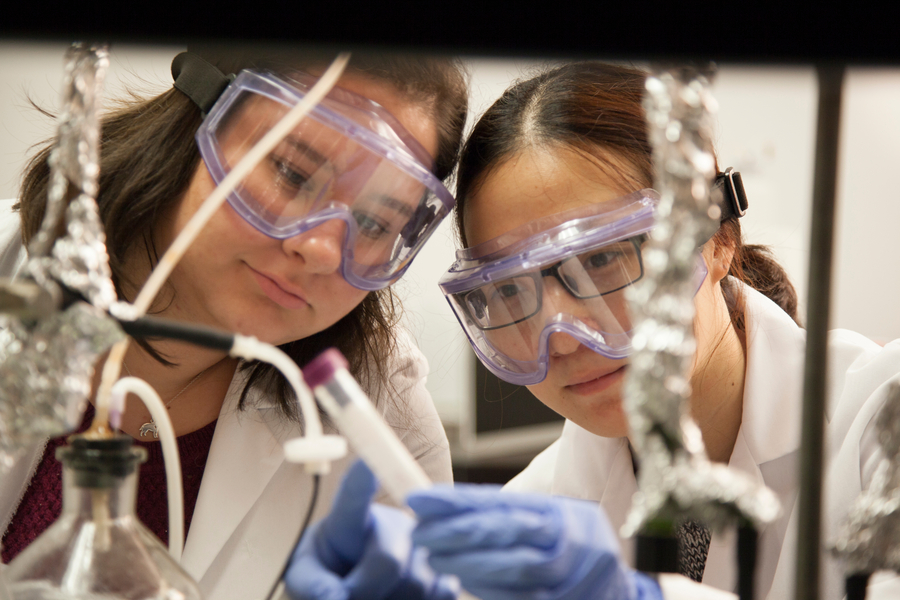 Two students wearing safety goggles and lab coats examine equipment closely during a chemistry experiment