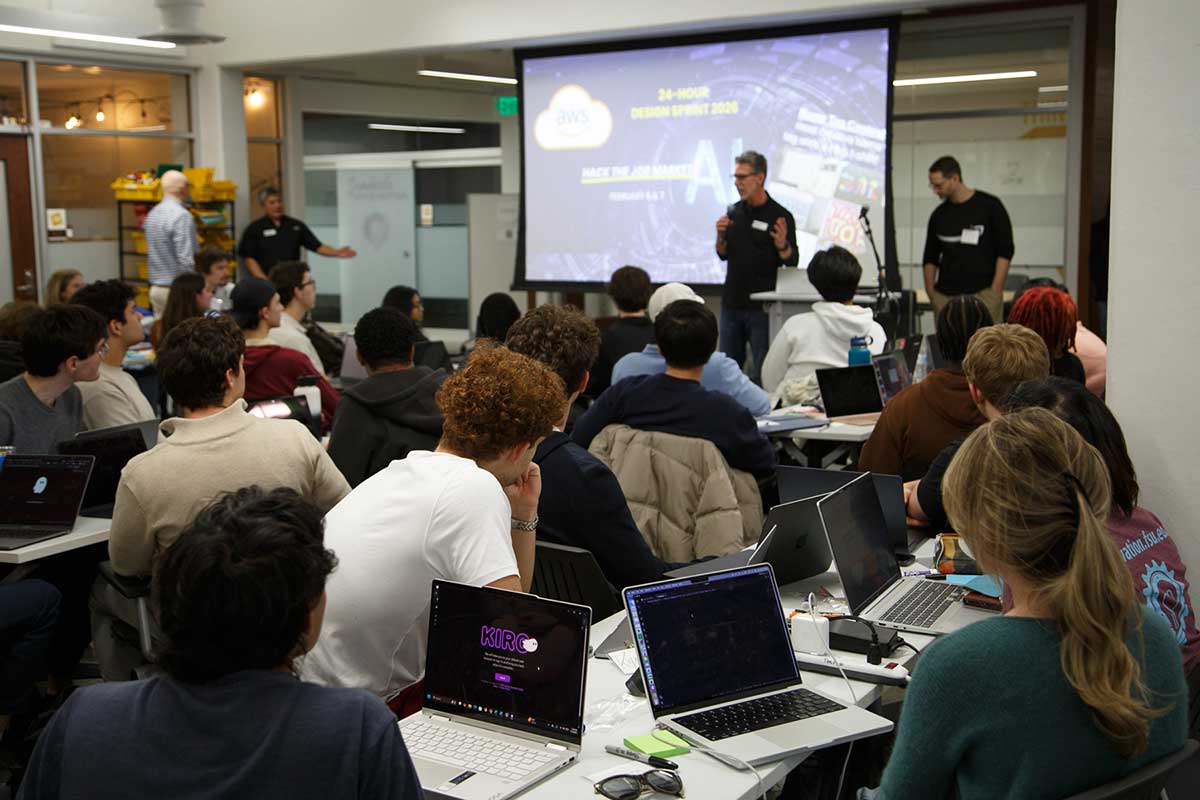 Presenters speaking to a room of students seated at laptops during an AWS design sprint event