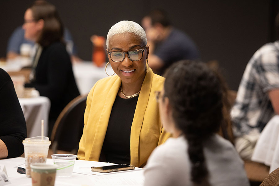 A faculty member in a yellow cardigan smiles while seated with colleagues at a table during a research workshop