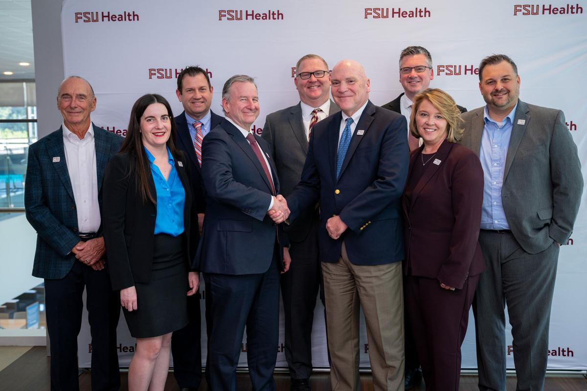 Representatives from Florida State University and Tallahassee Memorial HealthCare shake hands and pose in front of an FSU Health.