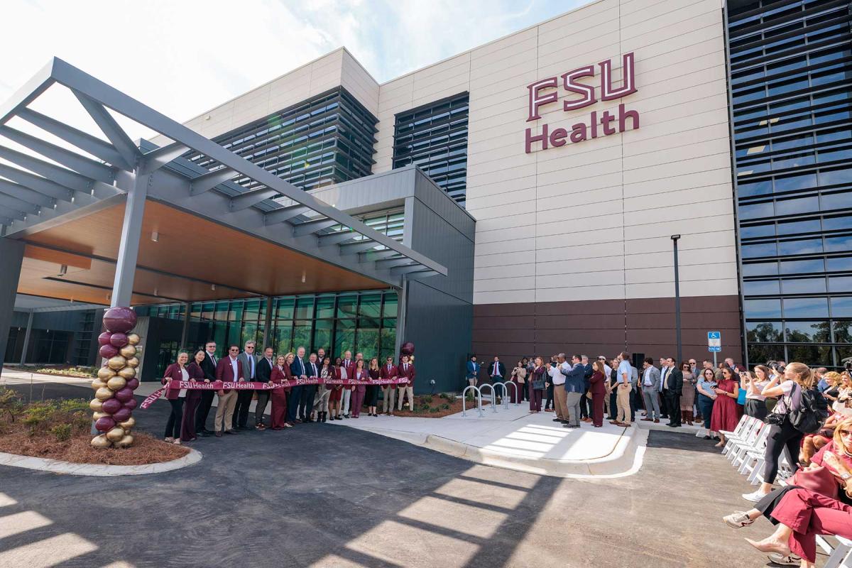 People gather outside the FSU Health building for a ribbon-cutting ceremony, with leaders holding a maroon ribbon in front of the entrance and spectators seated and standing nearby.