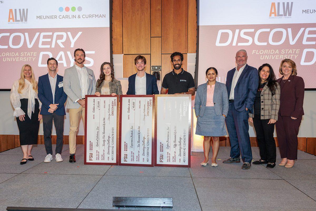 Ten people line up on a stage with Discovery Days shown on the screen. Three people in the middle are holding big checks.