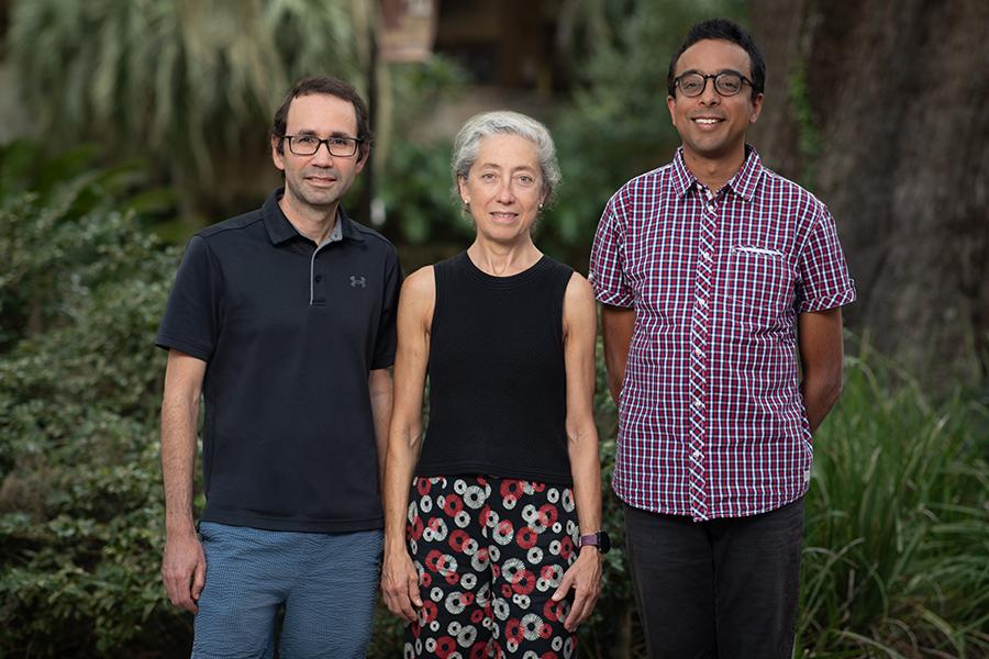 Three people stand side by side outdoors in front of greenery, smiling at the camera.