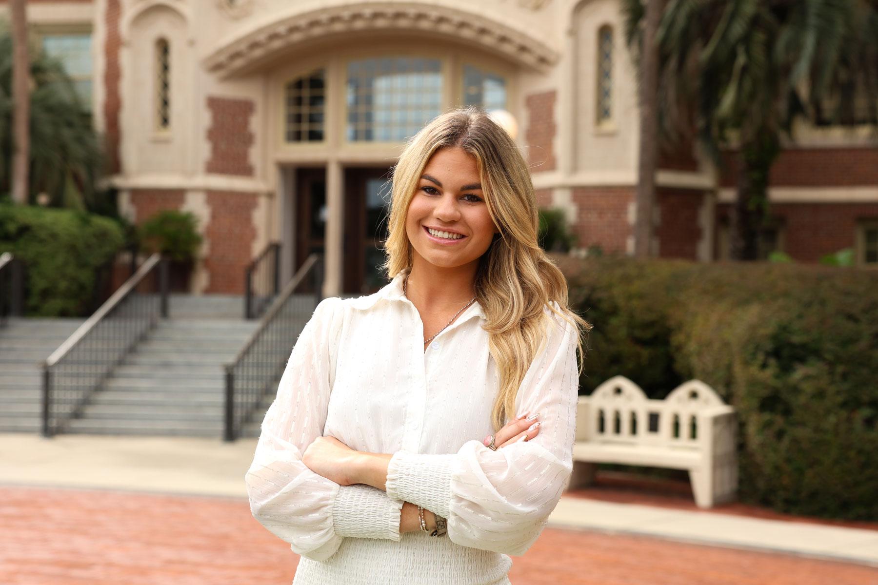 A smiling blonde woman stands with her arms crossed outdoors in front of a brick university building.